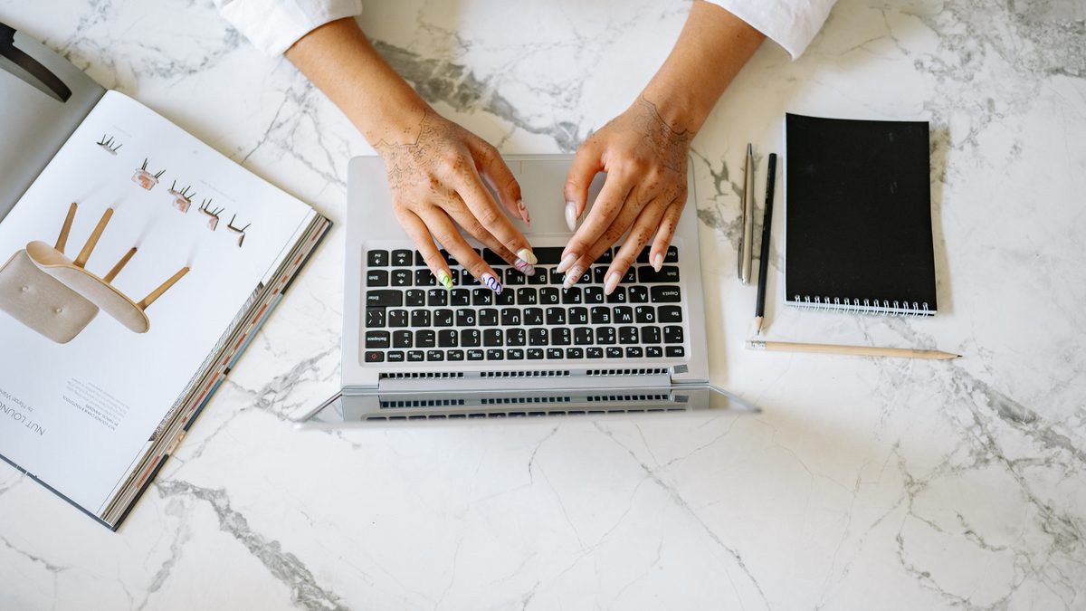 Overhead view of hands typing on a laptop at a marble desk beside a notebook and pens