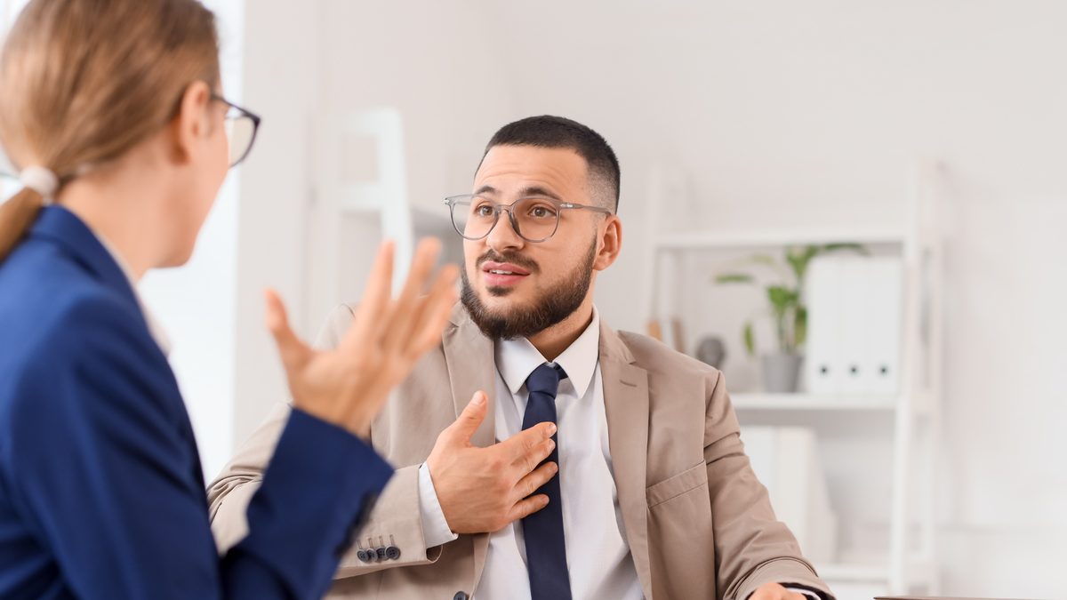 A businessman in glasses talking with a colleague across a desk in a bright office