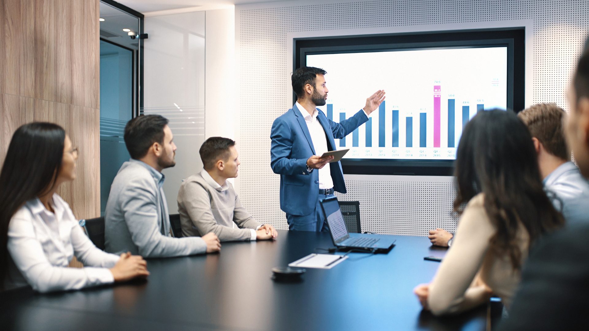 A boardroom team meeting around a table with a blank presentation screen