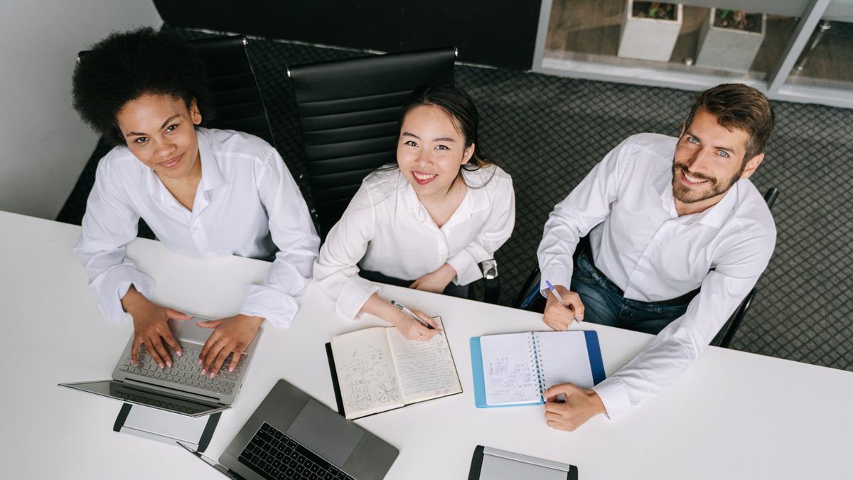 Three colleagues in white shirts collaborating at a table, taking notes and using a laptop
